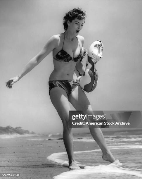 Low-angle portrait of an unidentified model, in a two-piece bathing suit, as she holds an inflatable seahorse under one arm and dips toes in the surf...