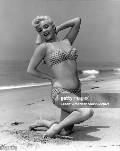 Portrait of an unidentified model, in a two-piece bathing suit, as she kneels in the sand on the beach, Los Angeles, California, January 25, 1958.