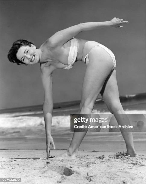 Portrait of an unidentified model, in a strapless, two-piece bathing suit, as she bends over to touch her toes at the beach, Los Angeles, California,...