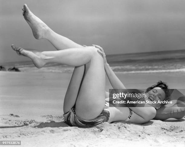 Portrait of an unidentified model, in a two-piece bathing suit, as she lies on her back, her legs in the air, on the sand at the beach, Los Angeles,...