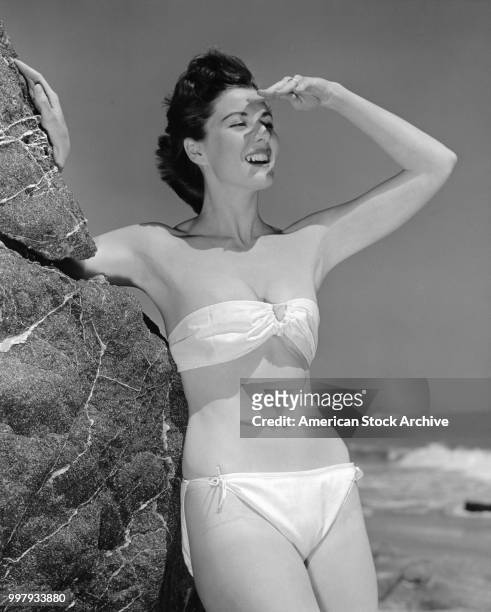 Portrait of an unidentified model, in a two-piece bathing suit, as she leans against a large rock using one hand to shield her face from the...