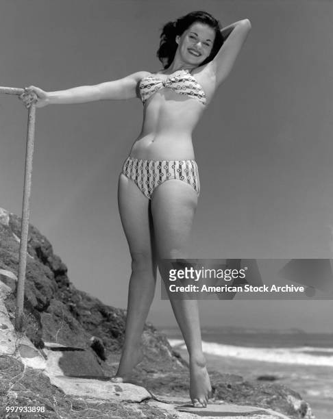 Low-angle portrait of an unidentified model, in a two-piece bathing suit, as she poses on a rocky outcrop at the beach, Los Angeles, California,...