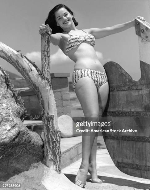 Portrait of an unidentified model, in a two-piece bathing suit, as she poses a beside a wooden shape at the beach, Los Angeles, California, January...