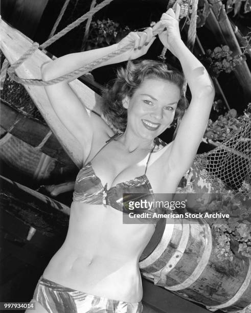 Portrait of an unidentified model, in a two-piece bathing suit, as she poses, hands above her hand holding into a rope, Los Angeles, California,...