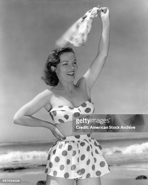 Portrait of an unidentified model, in a strapless, two-piece, polka-dot bathing suit, as she waves on the beach, Los Angeles, California, January 25,...