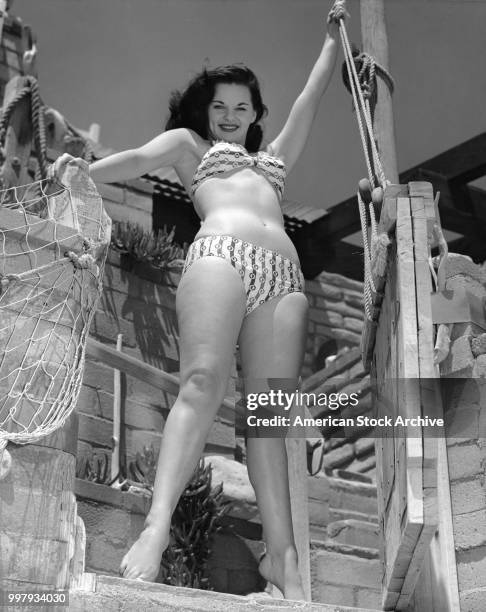 Low-angle portrait of an unidentified model, in a two-piece bathing suit, as she poses on a wooden structure draped with a fish net, Los Angeles,...