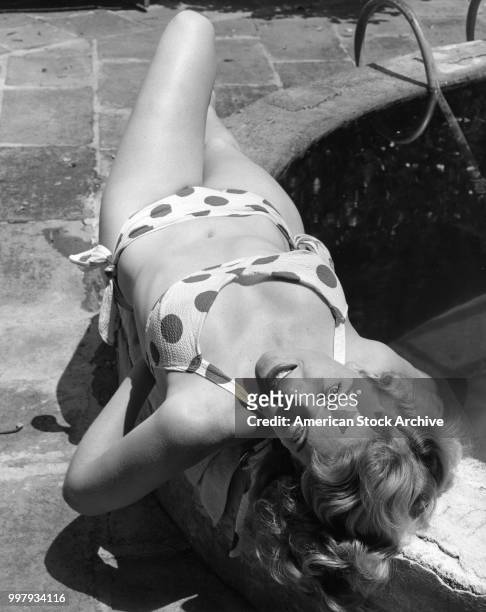 Portrait, from above, of an unidentified model, in a two-piece, polka-dot bathing suit, as she poses,on the on the low wall around a small pool in a...