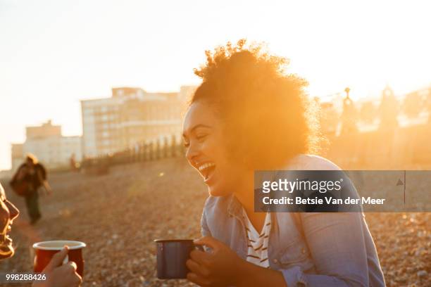 mixed race woman laughing with friend, having picnic on beach, backlit at dusk. - betsie van der meer stock pictures, royalty-free photos & images