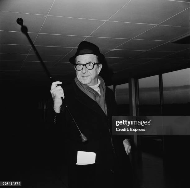 American comic and entertainer Phil Silvers waves a walking stick in the air as he walks along a corridor at Heathrow airport in London on 3rd...
