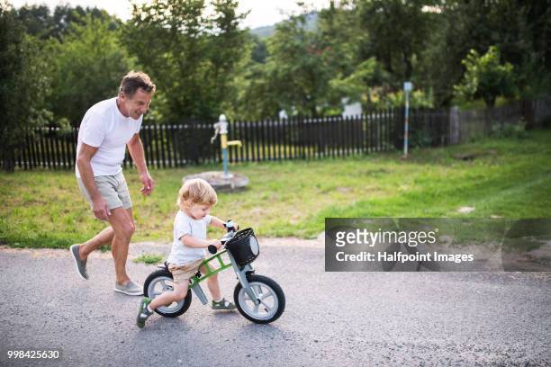 a happy small toddler boy with his grandfather riding a balance bike outdoors. copy space. - halfpoint stock-fotos und bilder