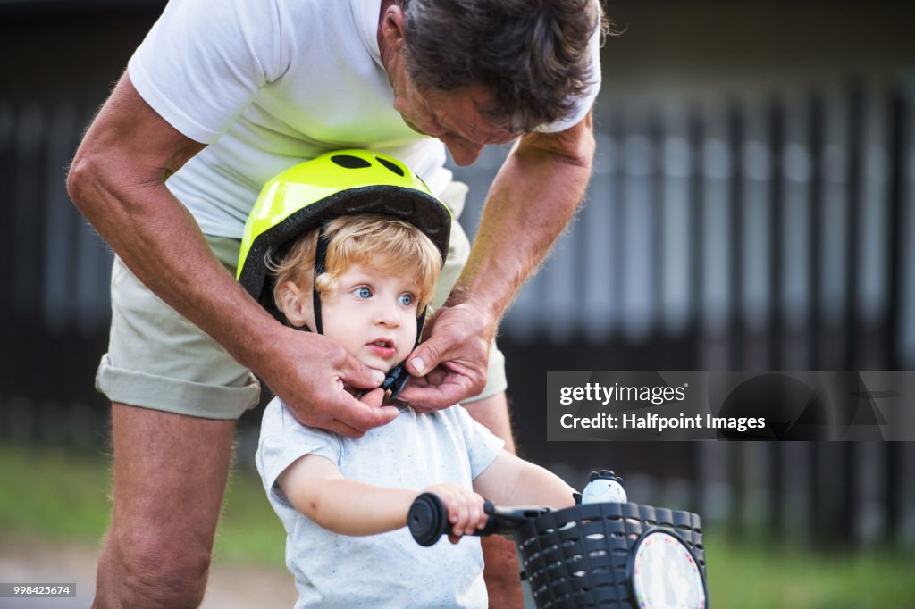 A senior grandfather putting a helmet on a small toddler boys head outdoors. Copy space.