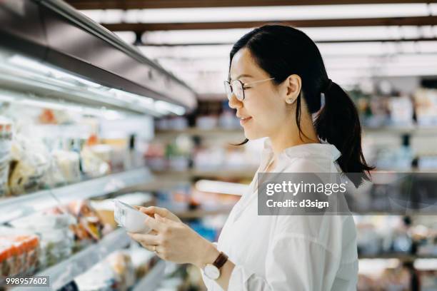 young asian woman grocery shopping in supermarket and reading nutrition label on a packet of fresh egg - fleischersatz stock-fotos und bilder