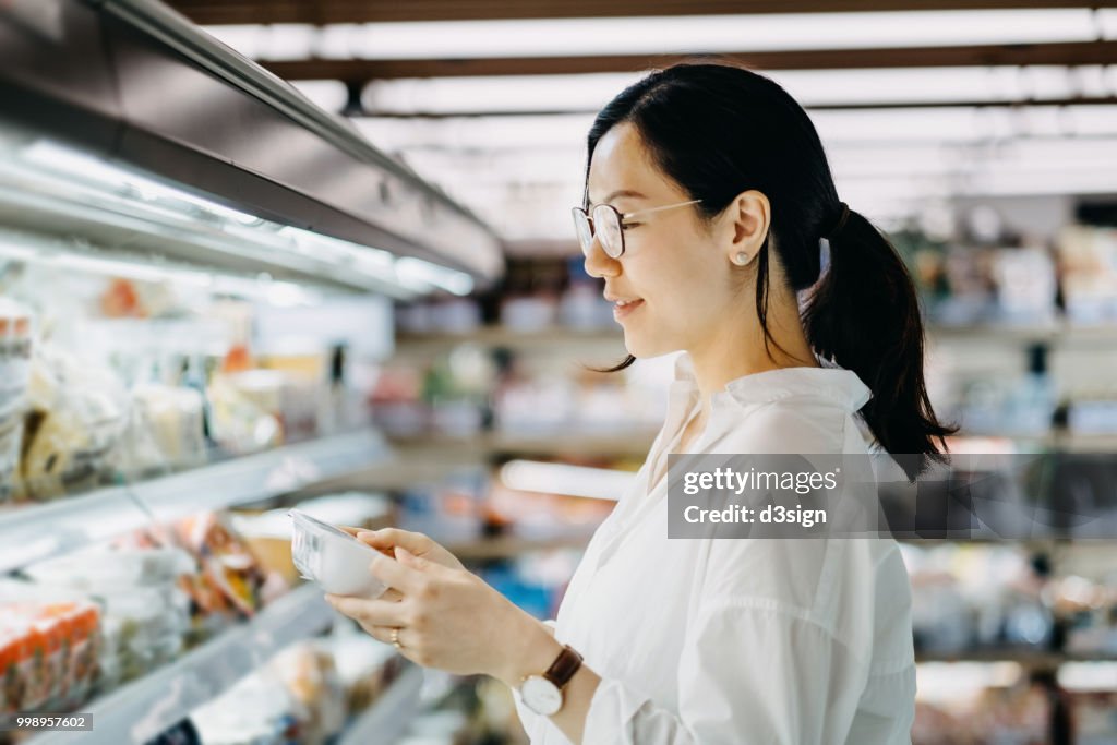 Young Asian woman grocery shopping in supermarket and reading nutrition label on a packet of fresh egg