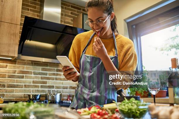 beautiful young woman using her smartphone while cooking - cooking cookbook stock pictures, royalty-free photos & images