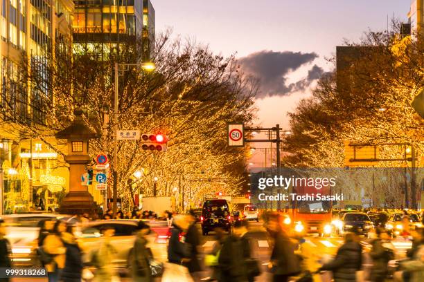 people walk and cars run on aoyama dori street, which street crosses the illuminated omotesando street at sunset at minamiaoyama, minato tokyo japan on december 05 2017. tree lined omotesando street is decorated and illuminated for winter holydays seasons - tree people stock-fotos und bilder