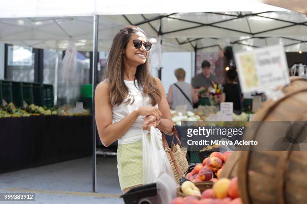 a young ethnic woman shopping at the farmers market, smiling, on a warm sunny day, wearing sunglasses. - mireya acierto stockfoto's en -beelden
