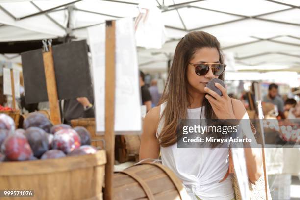 a young ethnic woman smells a plum, at the farmers market, on a warm sunny day, wearing sunglasses - mireya acierto stockfoto's en -beelden