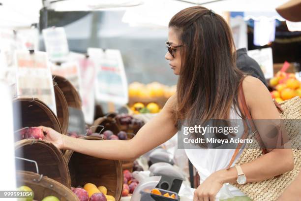 a young ethnic woman shopping for fruit at the farmers market, on a warm sunny day, wearing sunglasses - mireya acierto stockfoto's en -beelden