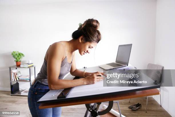 a young ethnic woman draws on her drafting table, with a laptop nearby, in her studio. - technophiler mensch stock-fotos und bilder
