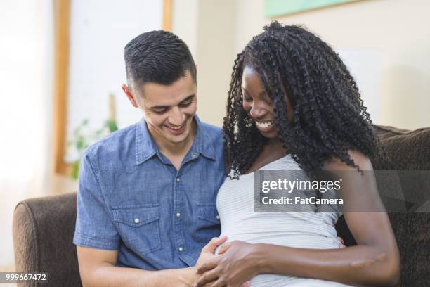 interracial pregnant couple snuggle on the couch together - maternity wear stock pictures, royalty-free photos & images