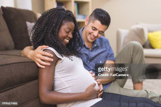 pregnant african american woman snuggles on the floor in front of the couch with her ethnic husband - maternity wear stock pictures, royalty-free photos & images