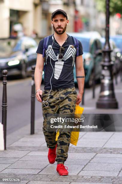 Guest wears a cap, a black t-shirt with a printed logo of the comics character Venom, green khaki military camo print pants, red sneakers, outside...
