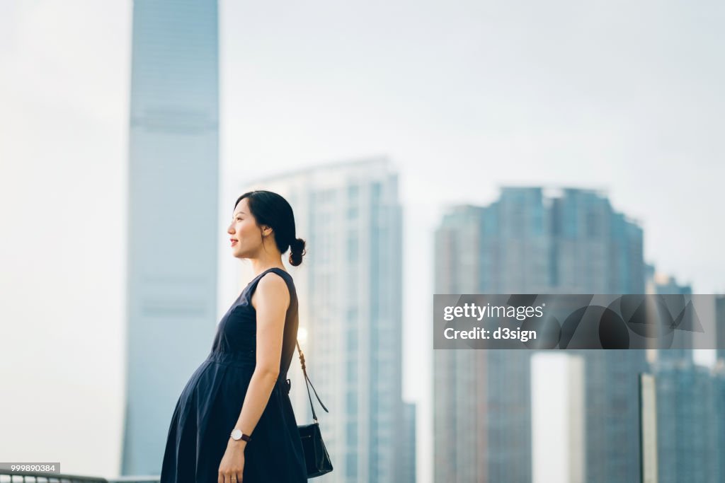 Beautiful pregnant Asian woman standing against modern cityscape enjoying view
