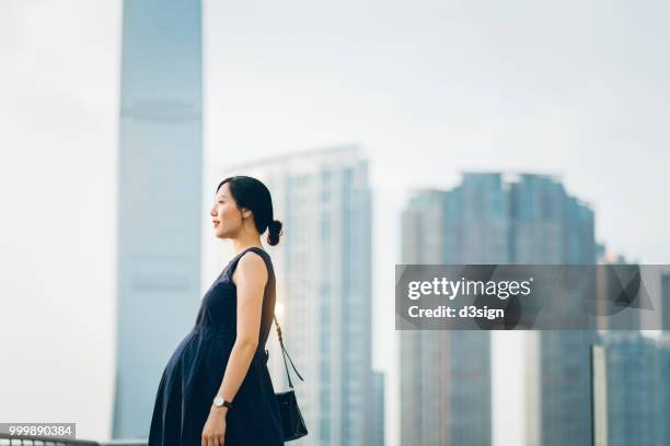 beautiful pregnant asian woman standing against modern cityscape enjoying view - new life sign stock pictures, royalty-free photos & images