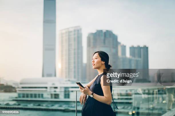 beautiful young pregnant woman relaxing on balcony using smartphone, standing against urban cityscape - asian woman pregnant stockfoto's en -beelden