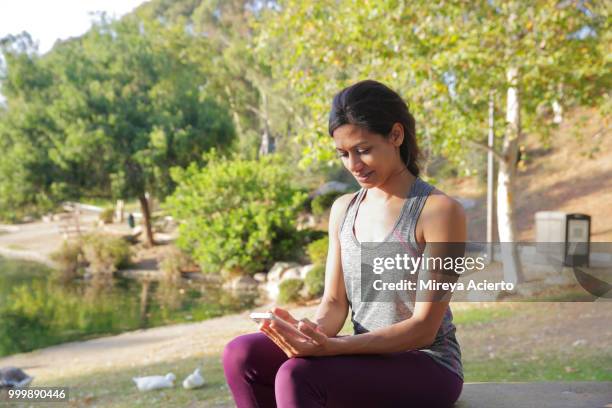 a fit, ethnic woman in athletic gear checks her phone while sitting on a picnic table in the park. - technophile stock pictures, royalty-free photos & images