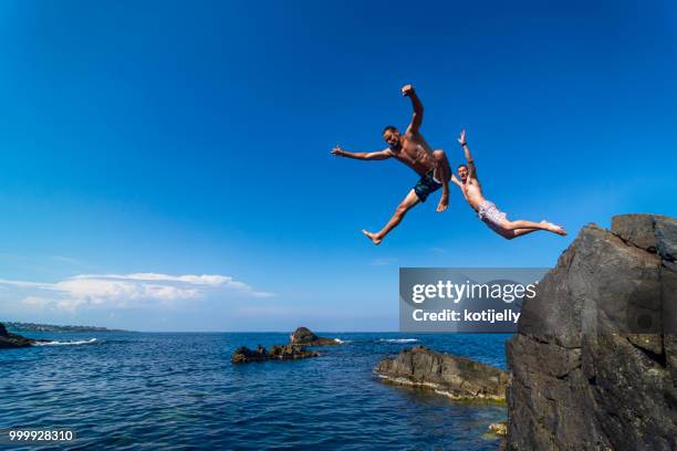 two young men jumping off cliff into sea - jumping sun stock pictures, royalty-free photos & images