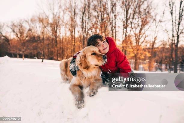 portrait of a boy sitting in the snow with his golden retriever dog - winter friends playing stock-fotos und bilder