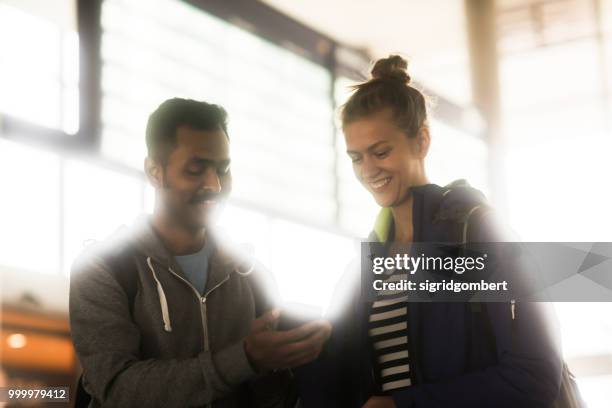 smiling couple standing in front of an arrival departure  board looking at a mobile phone - departure board front on stockfoto's en -beelden