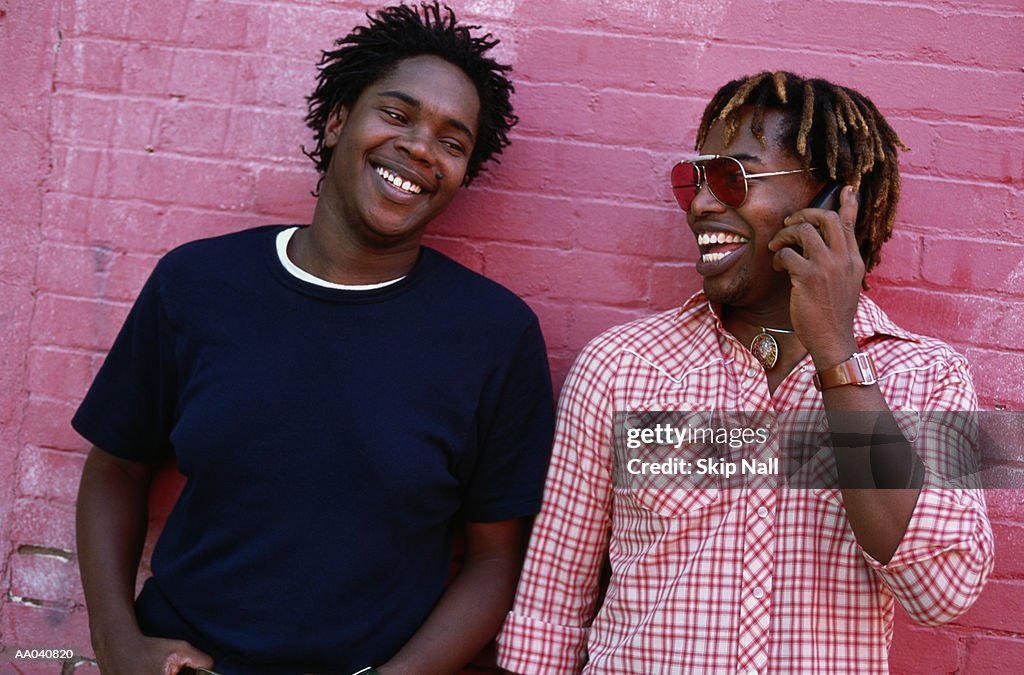 Two young men by pink brick wall smiling, one using mobile phone