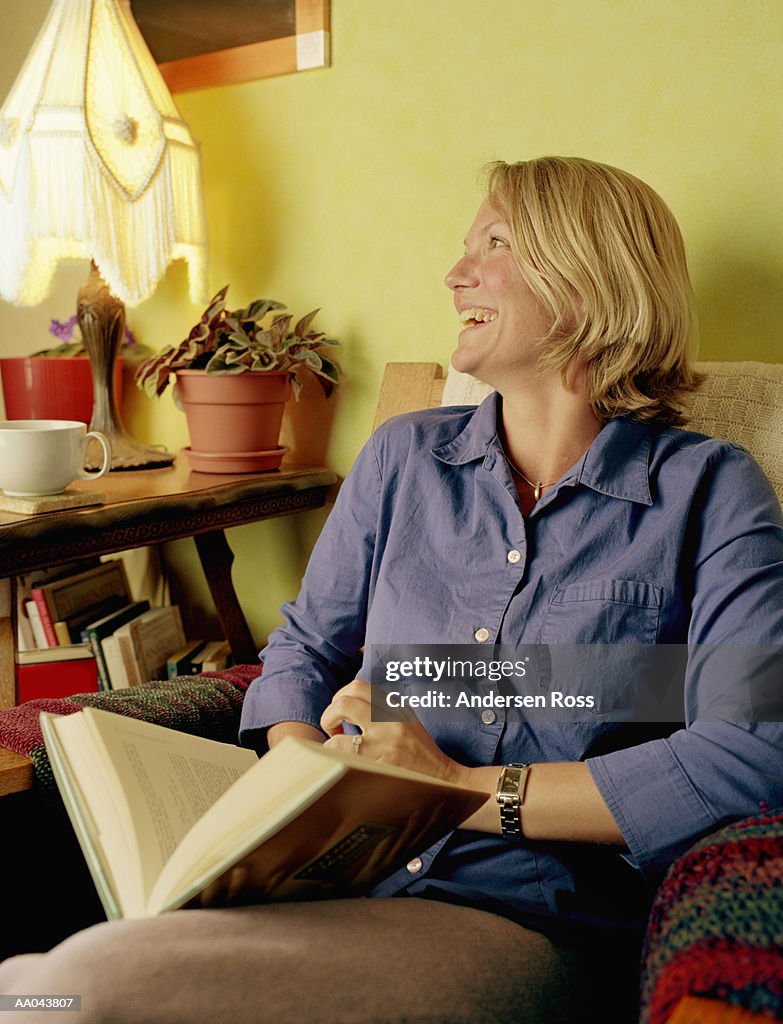 Woman sitting in armchair looking at book and laughing