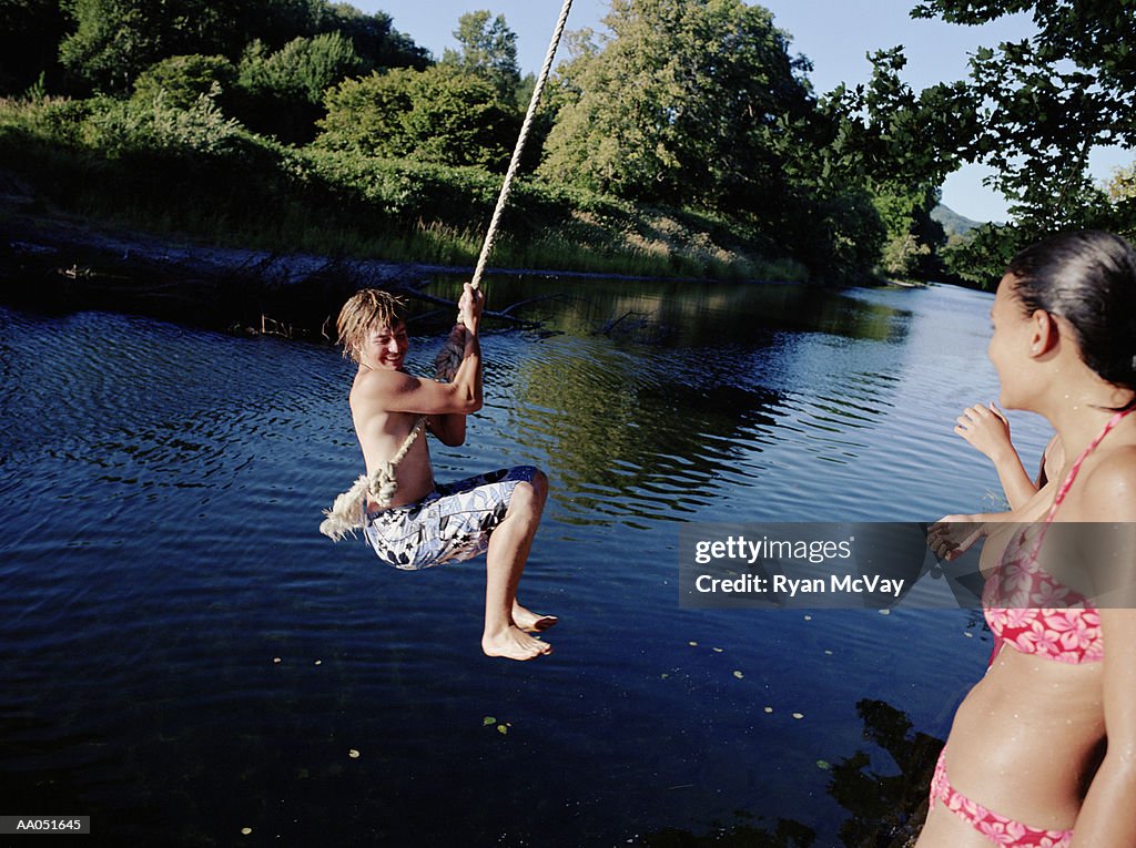 Teen girl watching teen boy (14-16) on rope swing over water, summer