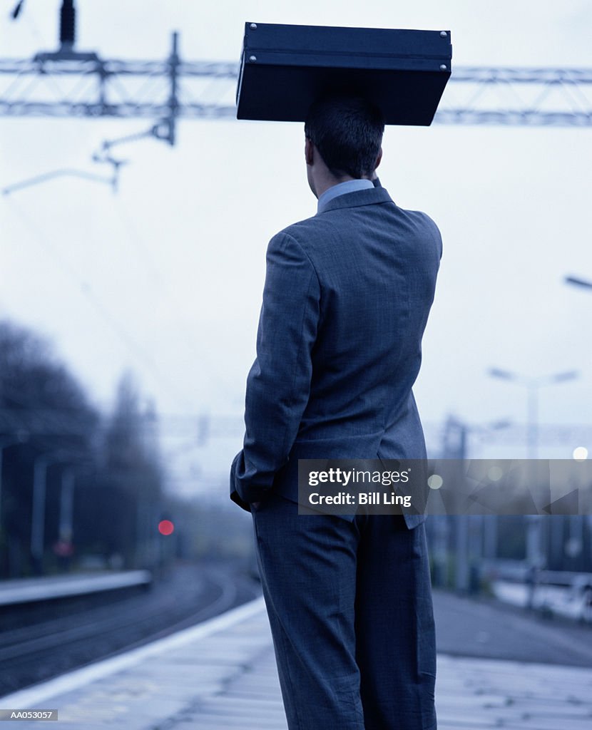 Businessman on train platform, using briefcase as umbrella, rear view