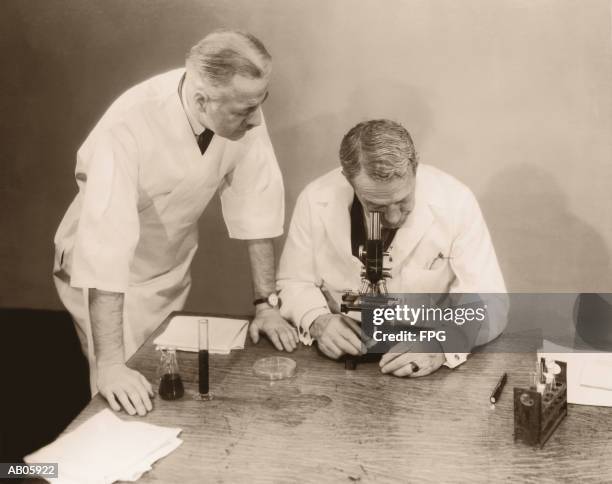 two men working in laboratory, one looking into microscope (b&w sepia) - 20th century stock pictures, royalty-free photos & images