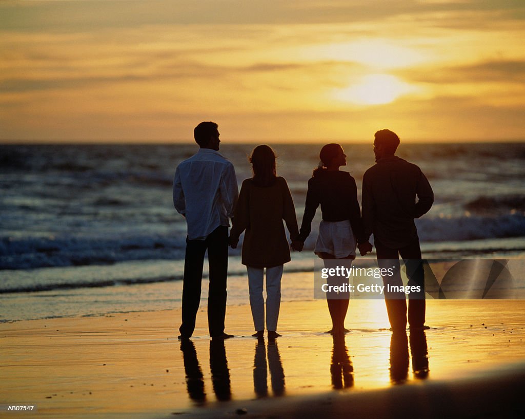 TWO COUPLES STANDING ON BEACH FACING SEA, AT SUNSET, PORTUGAL