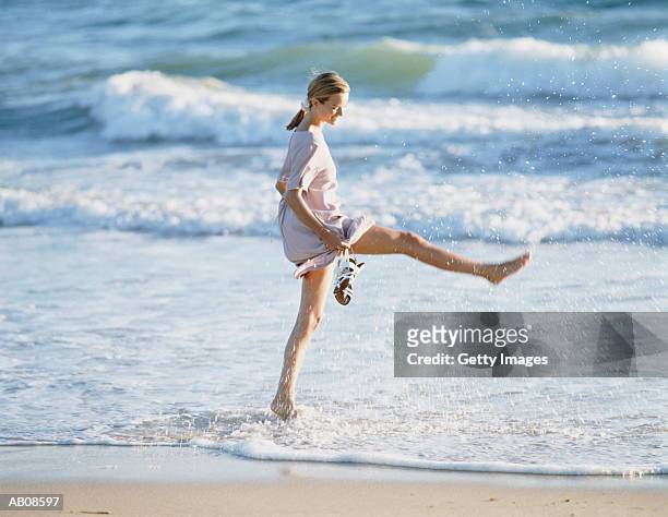 woman at water's edge on beach, holding hem of her dress up - knöcheltief im wasser stock-fotos und bilder