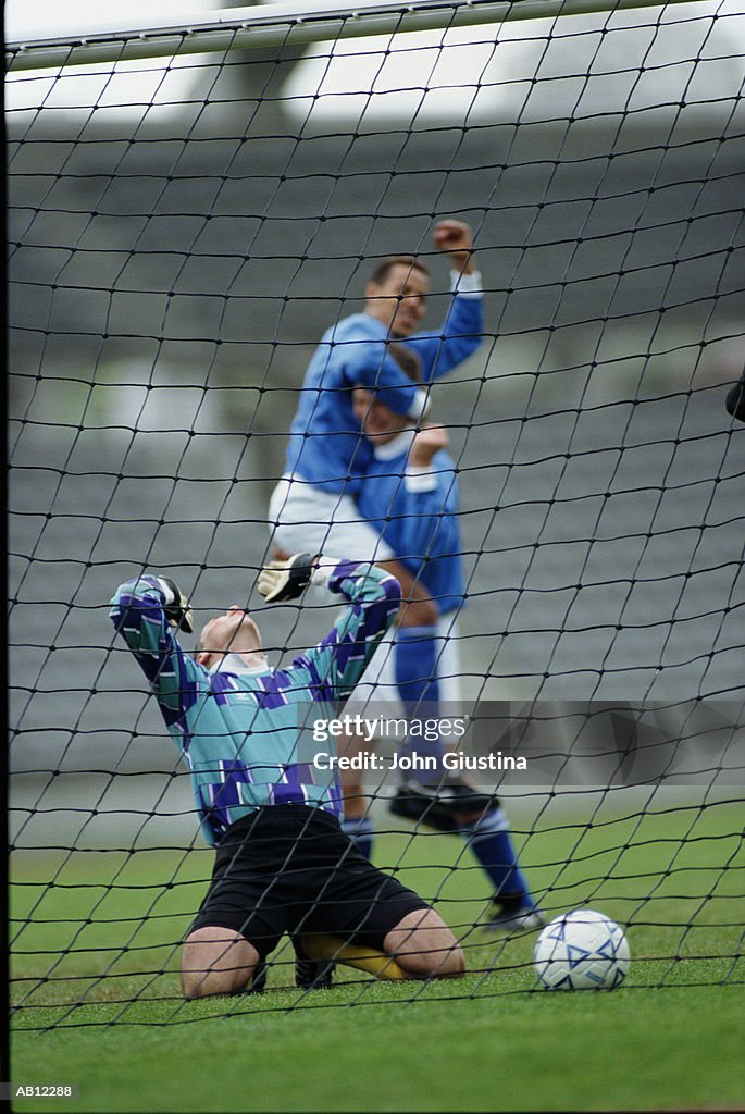 Goalkeeper kneeling in goal, opposing team celebrating score