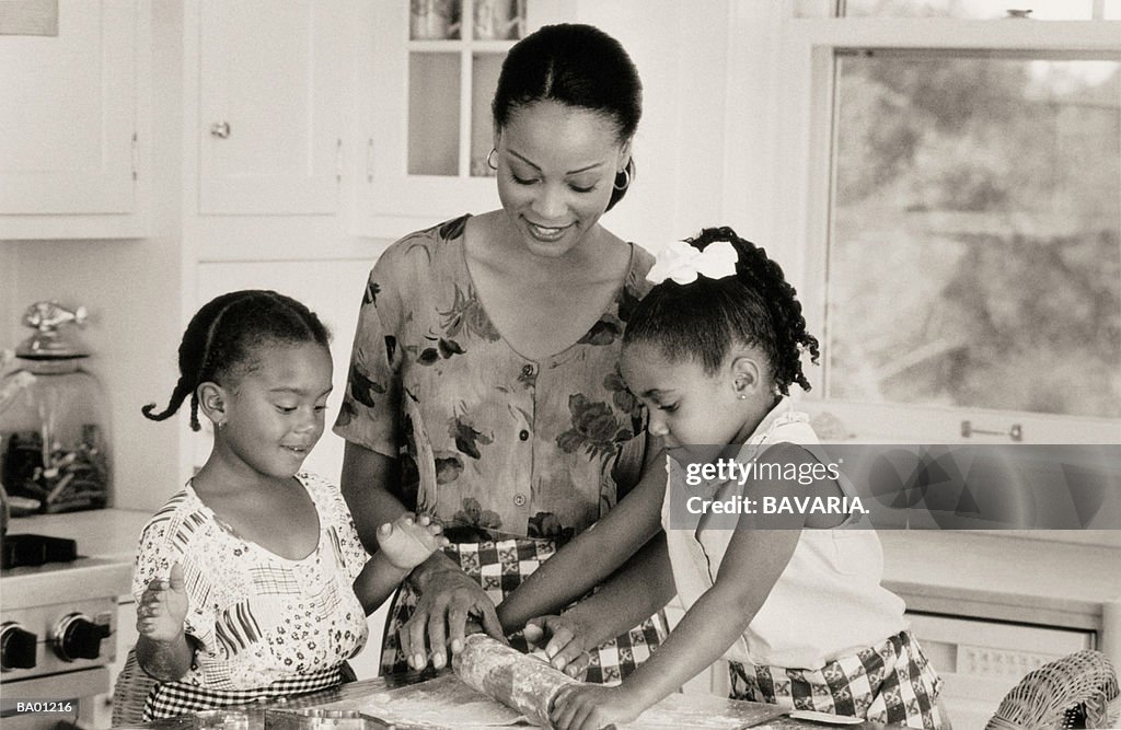 Mother and daughters (5-8) rolling pastry in kitchen (B&W)