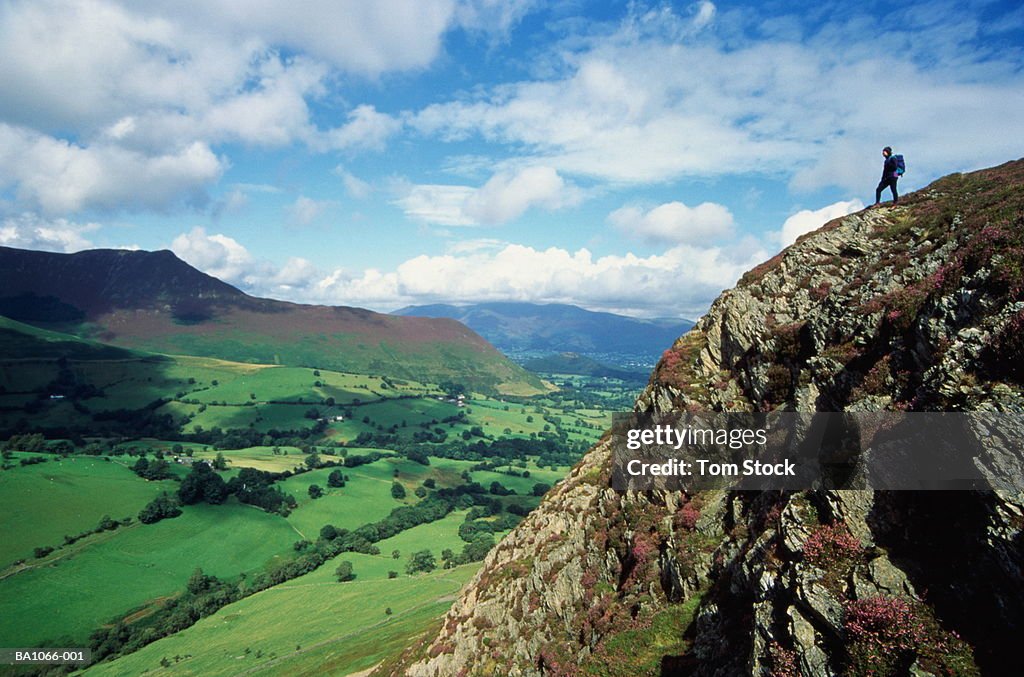 Walker standing atop high crag, overlooking verdant valley,England