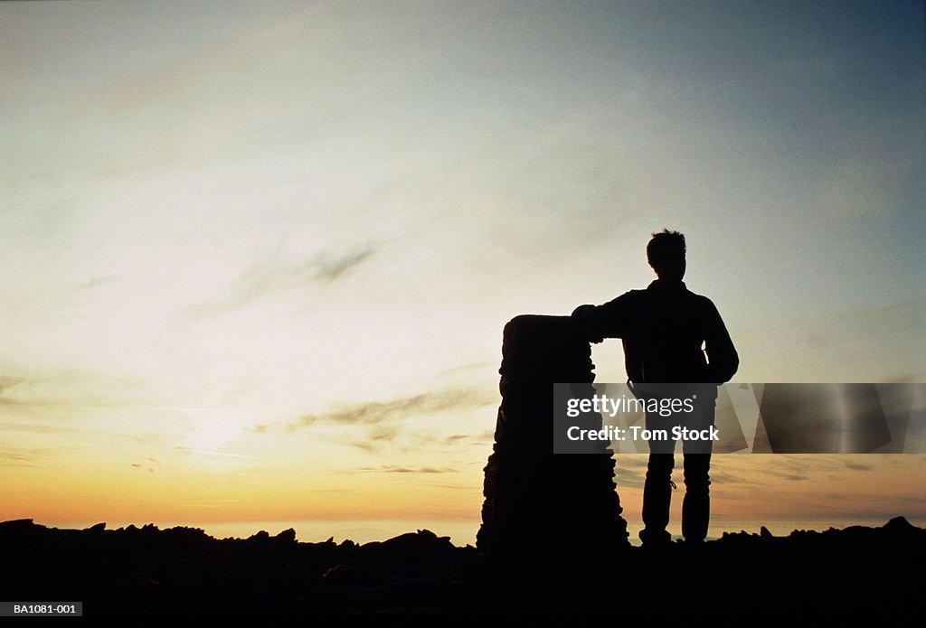 Hiker resting against cairn on hilltop, silhouette at sunset