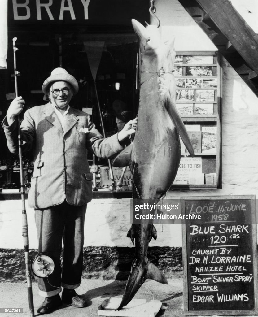 Fisherman standing by blue shark hanging on hook, portrait (B&W)