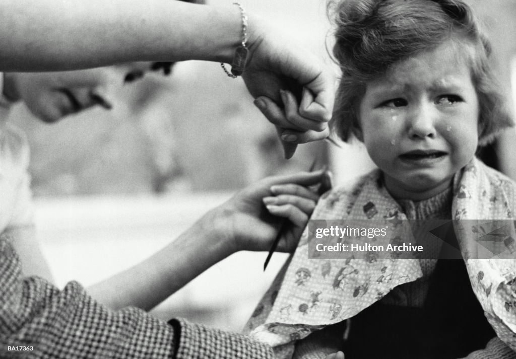 Hairdresser tending to crying girl's (3-5) hair, portrait (B&W)