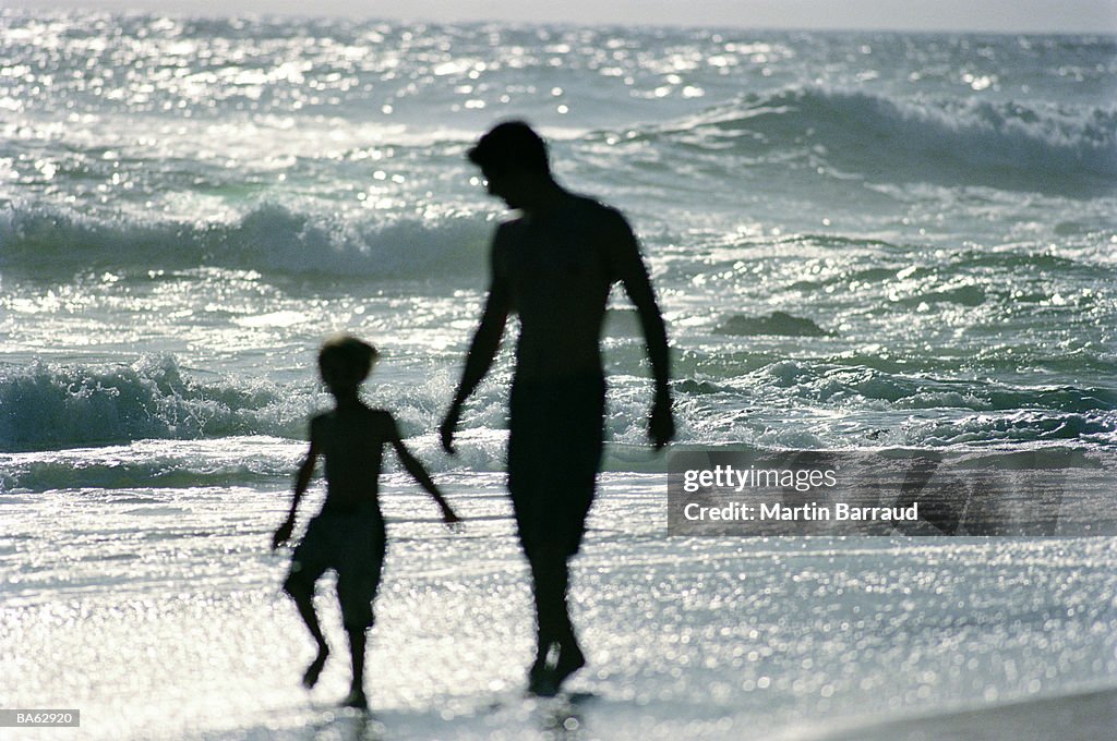 Father and son (4-6) walking in surf, silhouette