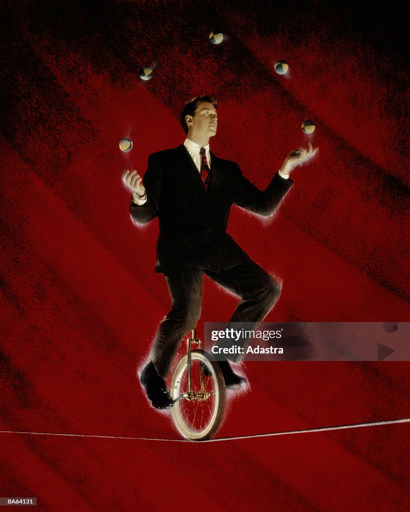 BUSINESSMAN ON ONE WHEELED CYCLE JUGGLING ON HIGH WIRE