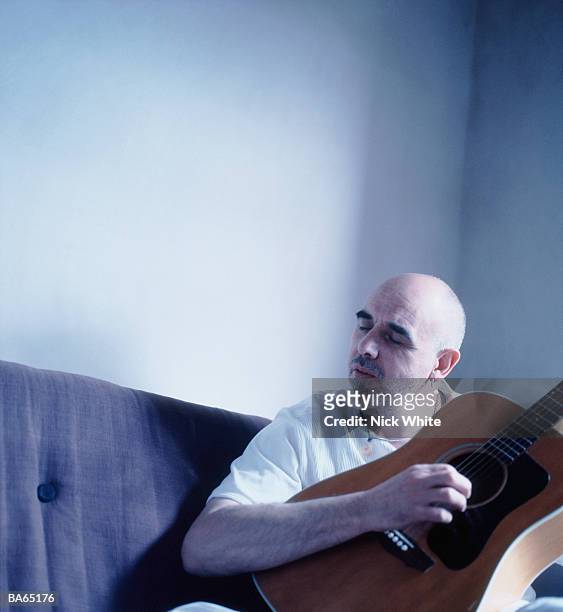 man sitting on sofa playing acoustic guitar, eyes closed - parcialmente calvo - fotografias e filmes do acervo