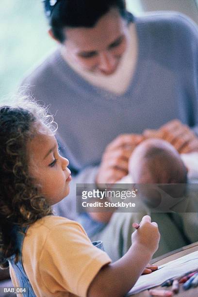 girl coloring at table, mother playing with baby in background - colouring stockfoto's en -beelden